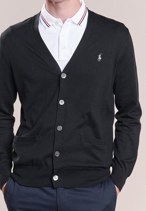 Mann trägt einen schwarzen, geknöpften Cardigan über einem weißen Poloshirt mit rot-blau gestreiftem Kragen, Hände in den Taschen.