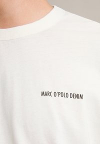 Balta t-kreklā, kas izgatavota no mīksta kokvilnas, ar apaļu izgriezumu un uz augšu drukātu melnu logo "MARC O'POLO DENIM".