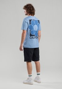 Light blue cotton t-shirt featuring a large, dark blue flower graphic on the back, paired with black shorts and white sneakers with green accents.