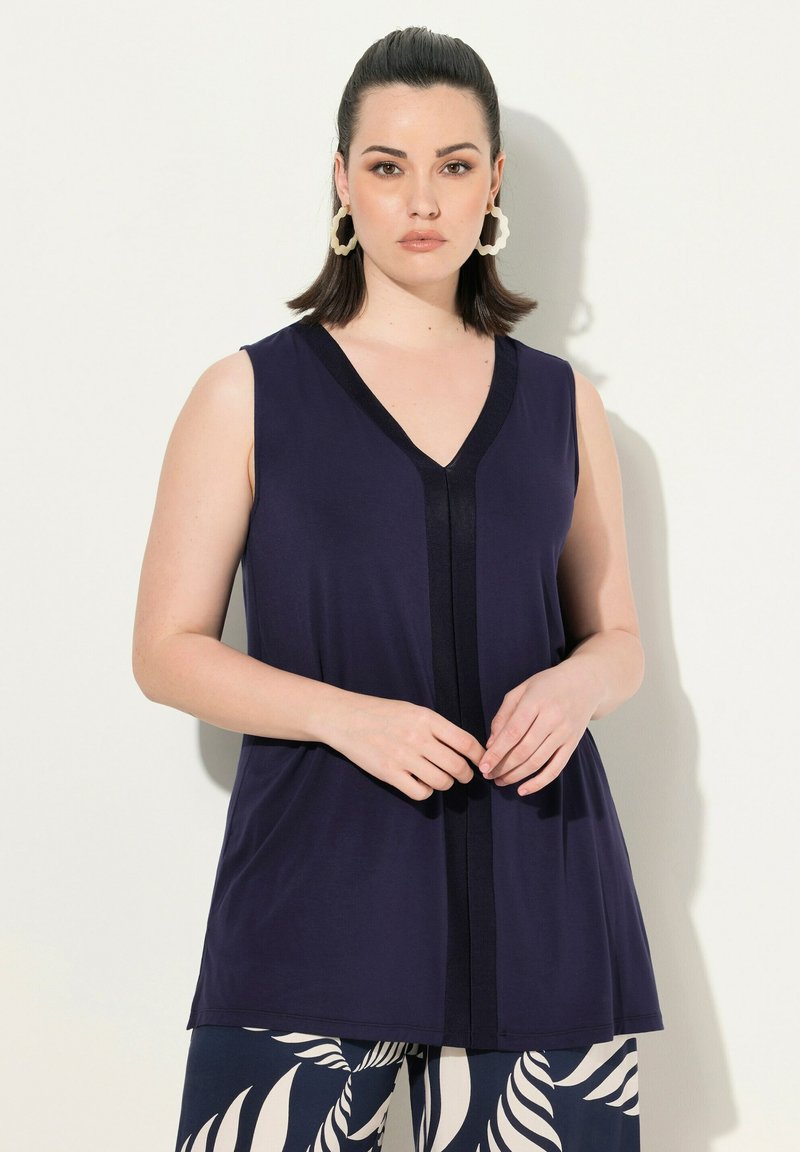 Marineblauwe mouwloze blouse met een V-hals en contrasterende zwarte accenten. Glad materiaal met een ontspannen pasvorm, gecombineerd met gestreepte broek.