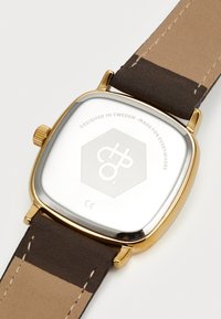 Reloj de cara cuadrada con la parte trasera de acero inoxidable pulido, caja en tono dorado y correa de cuero marrón bicolor. Presenta un logotipo grabado en la parte posterior.