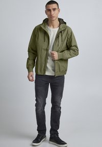 Giacca leggera color verde oliva con cappuccio, chiusura con zip, due tasche frontali e polsini elasticizzati, abbinata a una camicia chiara e jeans scuri.