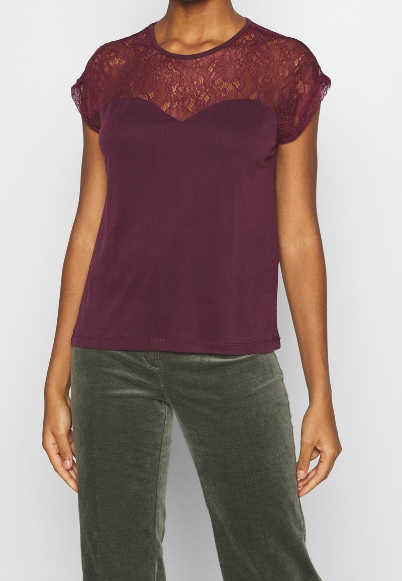 Femme portant un haut bordeaux manches courtes avec un empiècement en dentelle et un pantalon velours vert olive foncé, debout sur un fond clair.