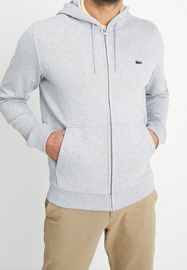Homme portant un sweat à capuche gris clair zippé avec des poches avant et un pantalon beige, mains dans les poches, debout devant un fond neutre.