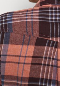 Chemise à carreaux dans des nuances de corail, marron et bleu ; tissu doux avec une texture lisse ; col ajusté et coutures visibles.