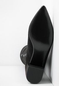 Sort ruskindsstøvle med høj hæl, spids tå, glat sort sål og tekstureret detalje langs skaftet. Enkel design, ingen synlige pynt.