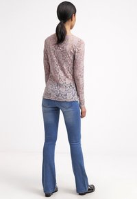 Top de encaje de manga larga en rosa claro con intrincados patrones florales, llevado sobre jeans acampanados azules, mostrando una textura semitransparente.