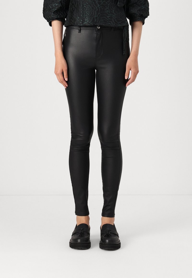 Femme portant un pantalon en cuir noir slim-fit avec fermeture par bouton et zip, associé à des mocassins noirs à pampilles sur fond blanc.