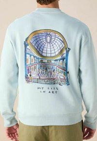 Sweatshirt bleu clair avec une illustration vibrante d'une galerie d'art au dos, agrémentée d'éléments décoratifs et du texte "MA VIE EN ART."