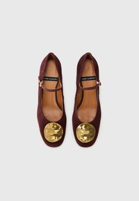 Ballerine in suede bordeaux con punta rotonda, dettagli in metallo dorato sul davanti e una sottile cinghia lungo il collo del piede.