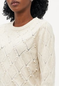 Kremo spalvos megztinis su deimanto raštu, papuoštas dekoratyvinėmis perlų detalėmis ir ažūrine struktūra. Klasikinis apvalus iškirpimas ir tekstūruotas paviršius.
