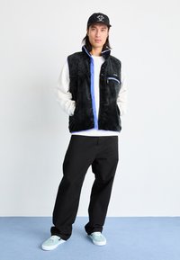 Black fleece vest with blue accents, zip pocket and button closure, worn over a white shirt and paired with black pants and mint sneakers.