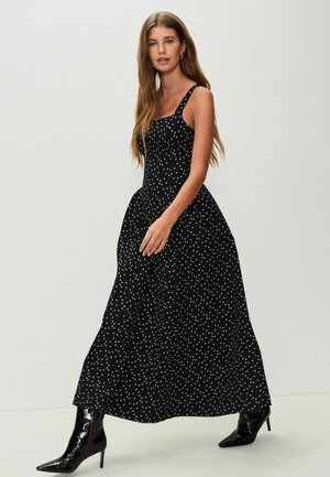Schwarzes Maxikleid mit weißen Polka-Dots, enganliegendem Oberteil und schwingendem Rock. Modell trägt schwarze glänzende spitz zulaufende Stiefel.