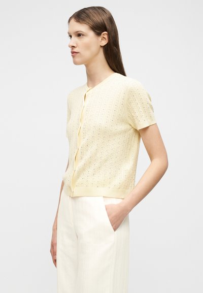 Γυναίκα που φοράει πλεκτή ζακέτα ανοιχτού κίτρινου χρώματος με κοντά μανίκια και λευκά παντελόνια ψηλής μέσης, στέκεται με το ένα χέρι στην τσέπη μπροστά σε απλό φόντο.