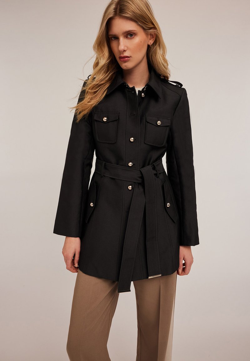 Abrigo trench negro con cinturón, cuatro bolsillos frontales, botones plateados y hombreras, combinado con pantalones rectos beige de pierna ancha.