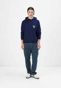 Marineblauwe hoodie met een voorzak, met witte tekst. Gekoeld met lichtblauwe spijkerbroek en witte sneakers met zwarte strepen.