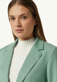 Mint green blazer with notched lapels, featuring a textured white sweater underneath. Notable details include a small logo accent on the lapel.