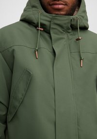 Hombre con chaqueta de invierno verde oliva con capucha y cordones con puntas de cobre, mostrando el torso frontal y la parte inferior de la cara.
