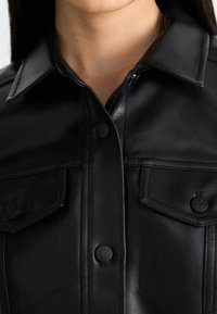 Zwart shirt van imitatieleer met knoopsluiting aan de voorkant, twee borstzakken en een kraag. Heeft subtiele logo-accenten op de knopen en de textuur.