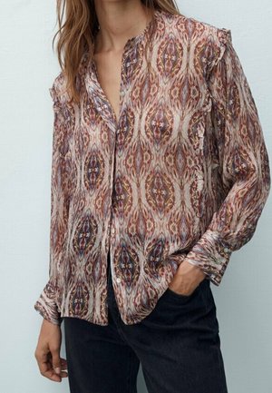 Blouse à manches longues avec boutons sur le devant, présentant un motif abstrait multicolore, portée avec un pantalon noir, une main dans la poche.