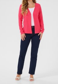 Roze cardigan met lange mouwen over een witte top, gecombineerd met donkerblauwe spijkerbroek en lichtgekleurde sandalen met hak. Gladde textuur en een losse pasvorm.