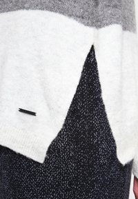 Hellgrauer und weißer Strickpullover mit dreieckigem Saumdesign, kombiniert mit dunkel strukturierten Hosen mit einem gesprenkelten Muster.