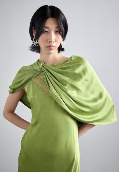 Donna con capelli neri corti, che indossa grandi orecchini di perle e un vestito di satin verde con dettagli in pizzo e una sciarpa drappeggiata, su uno sfondo grigio uniforme.