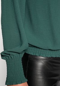 Grön långärmad blus med rynkade manschetter och textilstruktur, kombinerad med svarta läderbyxor, vilket framhäver kontrasterande material.