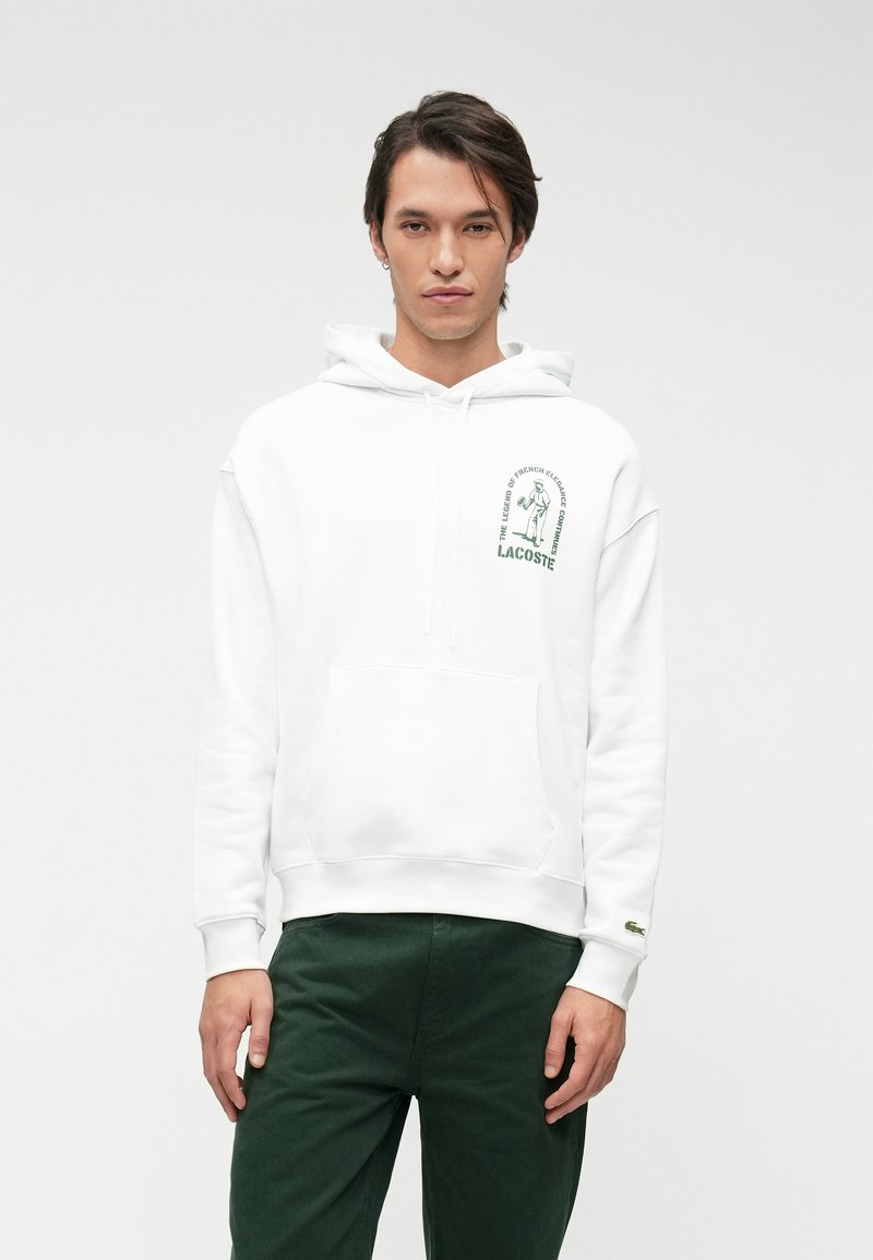 Hoodie blanc avec poche kangourou, capuche à cordon, et logo et texte Lacoste verts sur la poitrine. Porté avec un pantalon vert foncé.