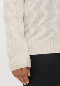 Pull en tricot torsadé couleur beige clair, avec un ourlet et des poignets côtelés, une texture douce et un design ajusté.