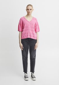 Ružový pletený kardigán s krátkymi rukávmi, V-výstrihom a gombíkovým zapínaním, s otvorenými vzormi. Skombinovaný s tmavo�šedými nohavicami a bielymi teniskami.