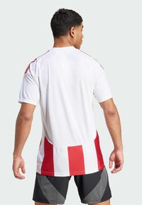 Witte sporttrui met rode strepen op de schouders en verticale rode strepen op de rug. Korte mouwen en een ronde halslijn.