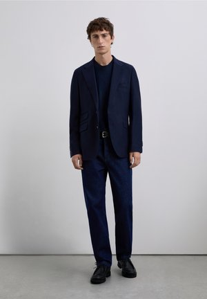 Blazer azul marino con solapas de muesca, dos bolsillos delanteros, llevado sobre una camiseta azul marino. Pantalones de mezclilla oscura y zapatos negros completan el conjunto.