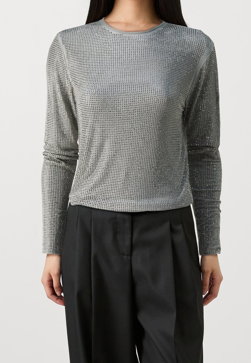Femme portant un haut argenté ajusté à manches longues avec un motif quadrillé texturé, associé à un pantalon noir large, sur un fond uni.