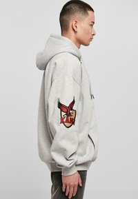 Sweatshirt à capuche gris avec une poche kangourou, arborant un logo d'aigle rouge brodé sur la manche gauche et une coupe décontractée.