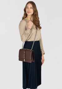Borsa a tracolla in pelle marrone con tracolla a catena, pattina pieghevole e logo inciso. La borsa è abbinata a una gonna blu a pieghe e a un top beige.