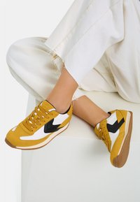 Gula och vita sneakers med svarta detaljer, med en låg design, texturerat mockamaterial och gummisulor.