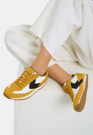 Zapatillas amarillas y blancas con acentos negros, que presentan un diseño de corte bajo, material de ante texturizado y suelas de goma.