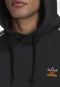 Svart hoodies med dragsnoddar, med en vit tre-stripesdesign på axlarna och en färgglad broderad Adidas-logga på bröstet.