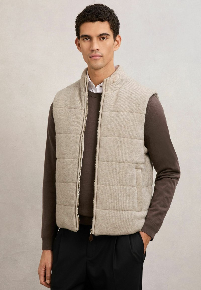 Mann trägt beige gesteppte ärmellose Weste über braunem Langarmshirt und schwarzer Hose, steht mit einer Hand in der Tasche vor schlichtem Hintergrund.