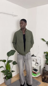 Olivgrön zip-up jacka med två bröstfickor, bärs över en svart skjorta. Ljust grå, avslappnad jeans och svarta stövlar. Inomhusmiljö med växter.