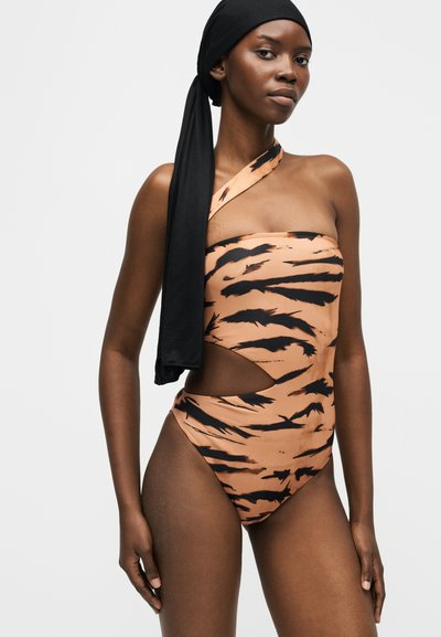 Γυναίκα με ολόσωμο μαγιό με ρίγες τίγρης σε έναν ώμο, με άνοιγμα στη μία πλευρά και μαύρο μαντήλι δεμένο στο πίσω μέρος.