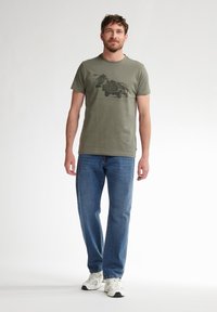 Olijfgroen t-shirt met een grafische print van een voertuig en natuur, gecombineerd met blauwe rechte jeans en witte sneakers.