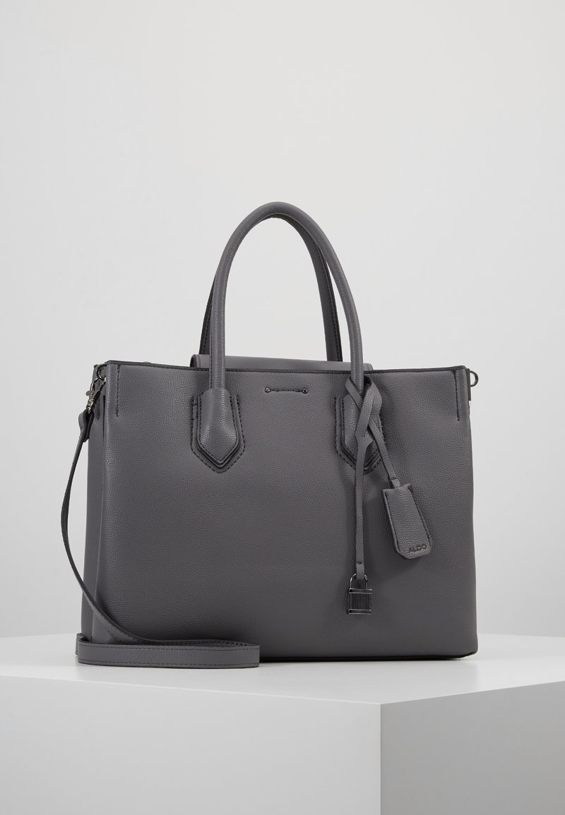 Sac cabas en cuir gris avec double poignée, une bandoulière amovible, et un verrou décoratif ainsi qu'une étiquette suspendus à l'avant.