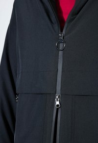 Černá bunda s vysokým límcem a dvojitým zipem; má texturovanou tkaninu a akcenty z kovových komponentů. Viditelný červený interiér.