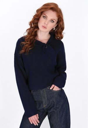 Navy niebieski krótki sweter z szerokimi rękawami i węzłem przy kołnierzu, zestawiony z ciemnoniebieskimi dżinsami. Gładka tekstura, swobodny fason.