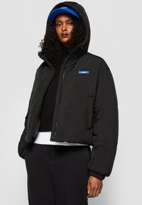 Melna cropped pūkainā jaka ar kapuci un rāvējslēdzēju priekšpusē, ar zilu zīmola etiķeti kreisajā krūtī. Modelis valkā melnu džemperi zem tās.