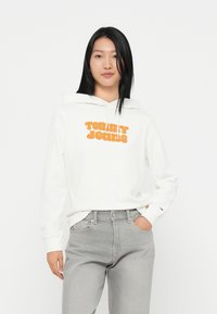 Felpa bianca con cappuccio e scritta arancione "Tommy Jeans". Presenta una tasca a marsupio e polsini a coste. Abbinata a jeans grigio chiaro.