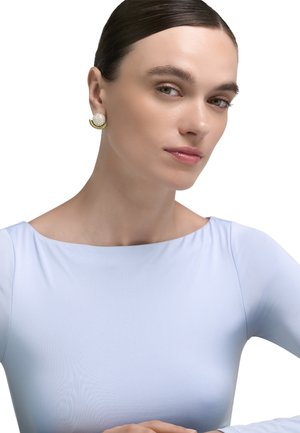 Naiste välimusega juuksed tagasikeeratud, seljas helesinine topp ja kuldsed kõrvarõngad pärlakaendustega, vaatab enesekindlalt kaamera suunas.