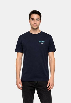 Jonge man draagt een marineblauw T-shirt met de tekst "PETROL" op de linkerborst, gecombineerd met zwarte jeans, staand tegen een effen achtergrond.